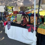 Outdoor market stall displaying a variety of colorful bags and fabrics on a black and white tablecloth under a canopy.