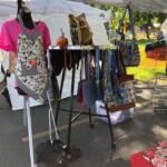 Outdoor market stall displaying aprons and bags on a rack under a tent.