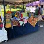 Table with colorful handmade quilts and fabrics displayed under a canopy at an outdoor market.