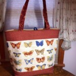 A tote bag with brown handles featuring a butterfly print design is placed on a surface in front of floral-patterned curtains.
