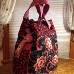 A floral-patterned fabric bag with red and pink flowers, has two handles and is placed on a wooden surface by a window.