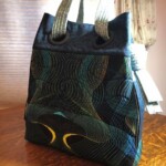 A multicolored, patterned tote bag with two handles sits on a wooden table near a window with curtains.