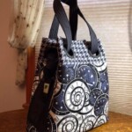A tote bag with black handles, featuring a pattern of clocks and houndstooth, sits on a wooden surface by a window.