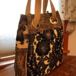 A large tote bag with blue floral pattern and beige straps is placed on a wooden table near a window with sheer curtains.