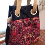 A patterned tote bag with large grommets and braided handles, featuring red, pink, and black floral designs, rests on a table.