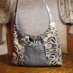 A patterned fabric handbag with a curved handle and button clasp is placed on a wooden table near floral curtains.