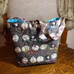 Gray quilted bag with sheep pattern on a wooden surface; blue interior and plaid straps; floral curtains in the background.