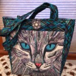 A patterned bag featuring a large cat face design with blue eyes, a blue and black geometric handle, and a decorative button.