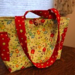 Colorful tote bag with insect and plant patterns, red star handles, a decorative flower button, on a wooden table.