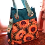 A colorful tote bag with sunflower prints and teal accents, featuring turquoise handles, placed on a wooden table.