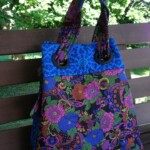 A colorful floral-patterned tote bag with circular metal handles rests on a wooden bench, set against a leafy green background.