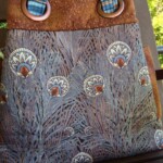 A tote bag with patterned fabric depicting peacock feathers and brown straps, hanging on a wooden chair.
