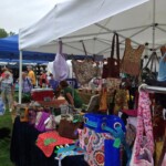 Outdoor market with various colorful bags hanging under a white canopy, and people browsing in the background.