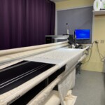 A long-arm quilting machine in a room with purple curtains and a computer monitor on the wall. Rolls of fabric are nearby.