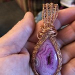 Hand holding a teardrop-shaped pendant with a pink geode encased in intricate copper wire wrapping.