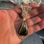 Hand holding a wire-wrapped pendant with a striped stone, displayed on a background of jeans and crafting supplies.