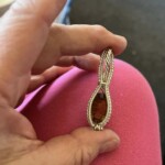 A hand holds a handcrafted metal wire pendant with an orange-brown stone, against a background of pink fabric.