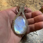 Teardrop moonstone pendant with intricate wire wrapping and a small red gemstone, held in hand at Creative Craftworks.