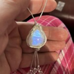 Hand holding a teardrop-shaped blue gemstone pendant with wire wrapping and dangling beads against pink plaid fabric at Creative Craftworks