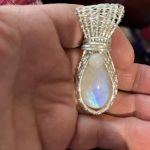 Hand holding a teardrop-shaped pendant with a silver wire wrap and an iridescent stone.