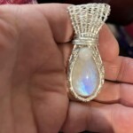 Hand holding a teardrop-shaped pendant with a silver wire wrap and an iridescent stone.