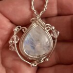 Close-up of a hand holding a teardrop-shaped pendant with a translucent stone encased in intricate silver wire wrapping.