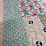 Close-up of a quilt with geometric and floral patterns in shades of blue, pink, and white, featuring detailed stitching.